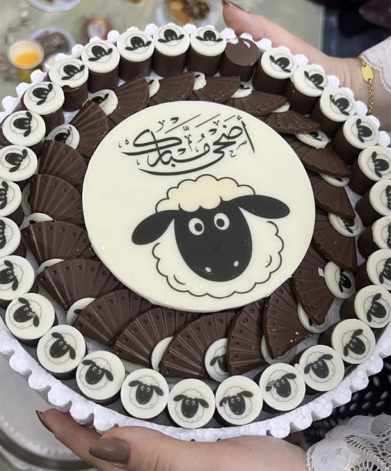 صينية دائرية كبيرة محاطة بحبّات شوكولاتة بيضاء مطبوع عليها وجه خروف، وفي وسطها قرص أبيض مكتوب عليه «أضحى مبارك» بخط عربي مع رسم خروف، وحولها قطع شوكولاتة داكنة مضلّعة.
