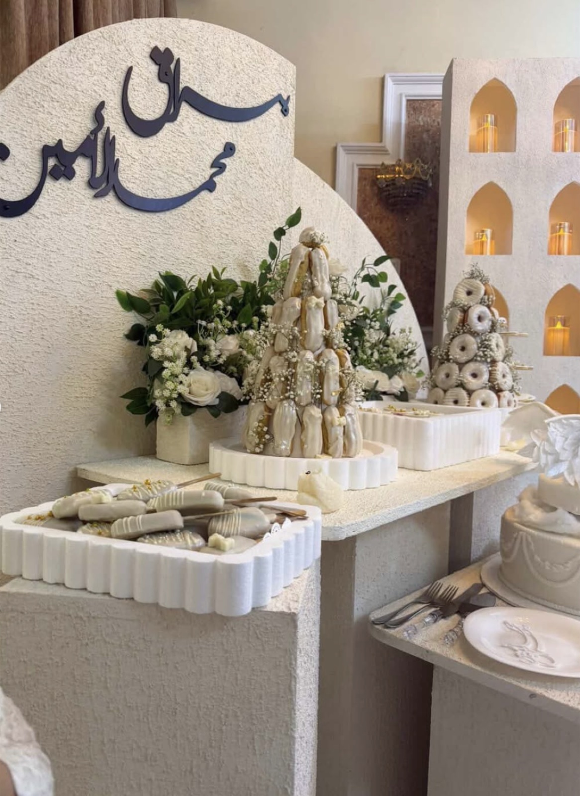 ركن حلويات أمام خلفية قوسية بيضاء تحمل اسمَي العروسين بخط عربي: برج فراولة مغطّاة بالشوكولاتة البيضاء، وبرج دونات، وصواني حلوى بيضاء مضلّعة، وورد أبيض وأخضر، وكيك طبقات.