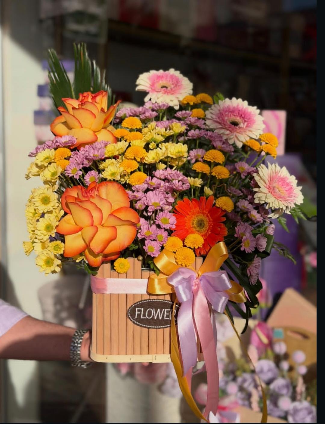سلّة خشبية مكتوب عليها FLOWER تحمل ورداً برتقالياً وجربيرا وردية وبيضاء وأقحواناً أصفر وبنفسجياً، مربوطة بشرائط وردية وذهبية.