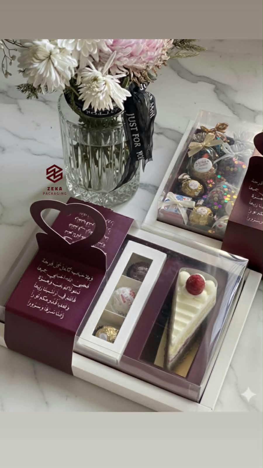 علبة عنابية بغطاء شفاف فيها ثلاث حبّات شوكولاتة وقطعة كيك مثلّثة بحبّة توت، وبطاقة قلب مطبوعة بشعر عربي، وخلفها علبة شوكولاتة ملوّنة ومزهرية ورد بشريط أسود.