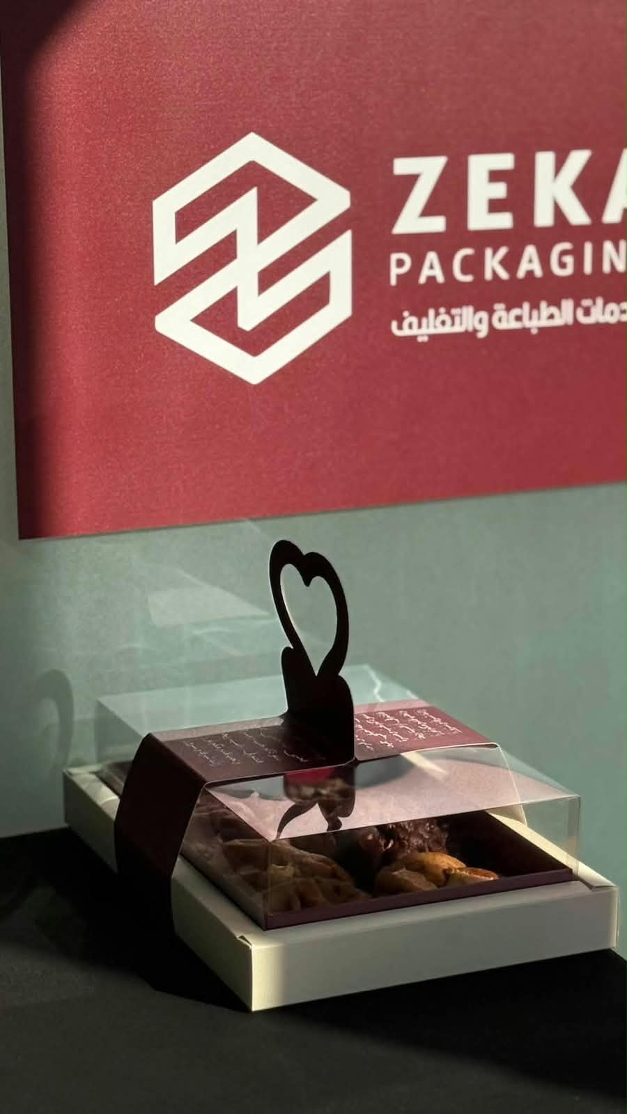 علبة بيضاء بغطاء شفاف وبطاقة عنابية بقلب مقصوص، أمام لافتة المحلّ العنابية المكتوب عليها ZEKA PACKAGING، خدمات الطباعة والتغليف.